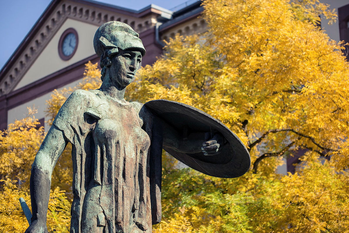 Bronzeenstatue einer Kriegerin mit Helm und Schild vor gelbem Herbstlaub.