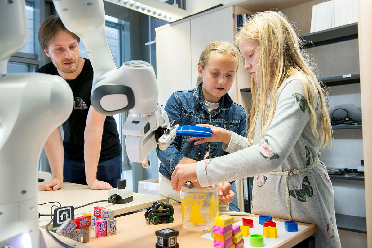 Zwei Mädchen und Wissenschaftler arbeiten mit einem Roboter; (c) KIT