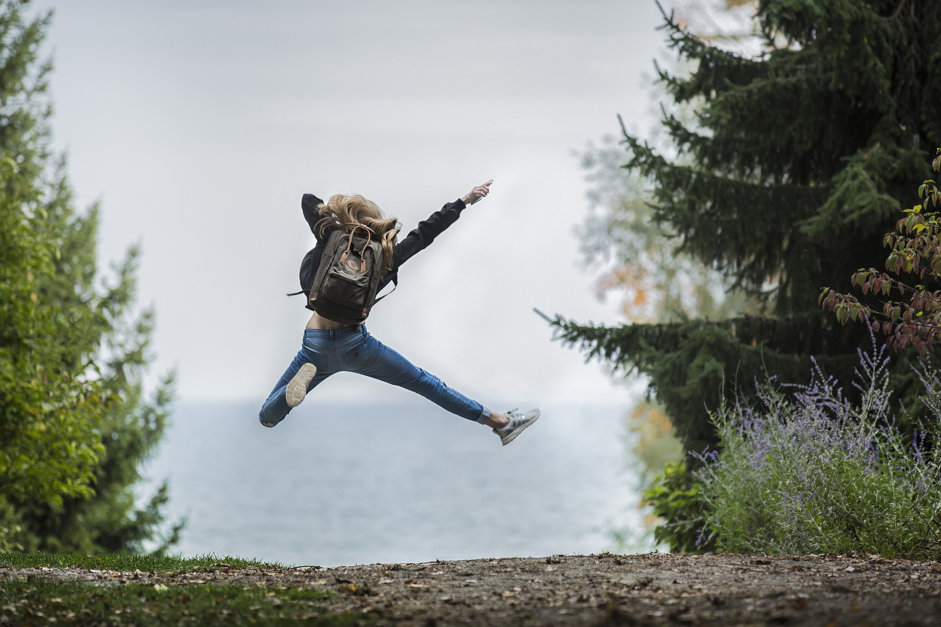 Foto von hinten: Junge Frau macht einen Luftsprung, vor sich das Meer, hinter sich ein steiniger Weg